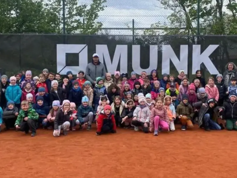 Große Kindergruppe beim Schnuppertennis auf dem Sandplatz vor dem Banner des Sponsors Munk